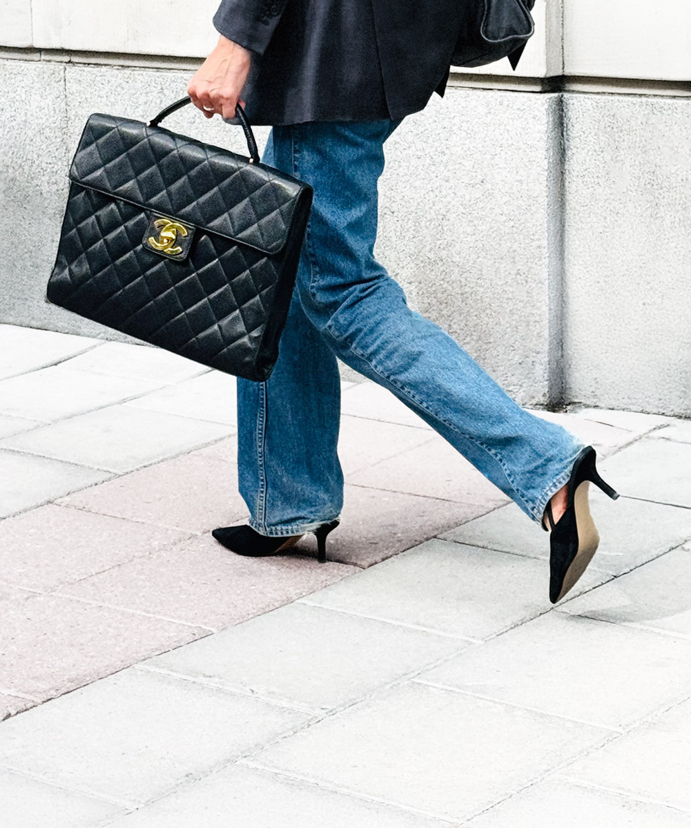 A woman in a dark blazer, jeans, and heels walking with a black quilted Chanel handbag featuring gold hardware, exuding timeless sophistication.