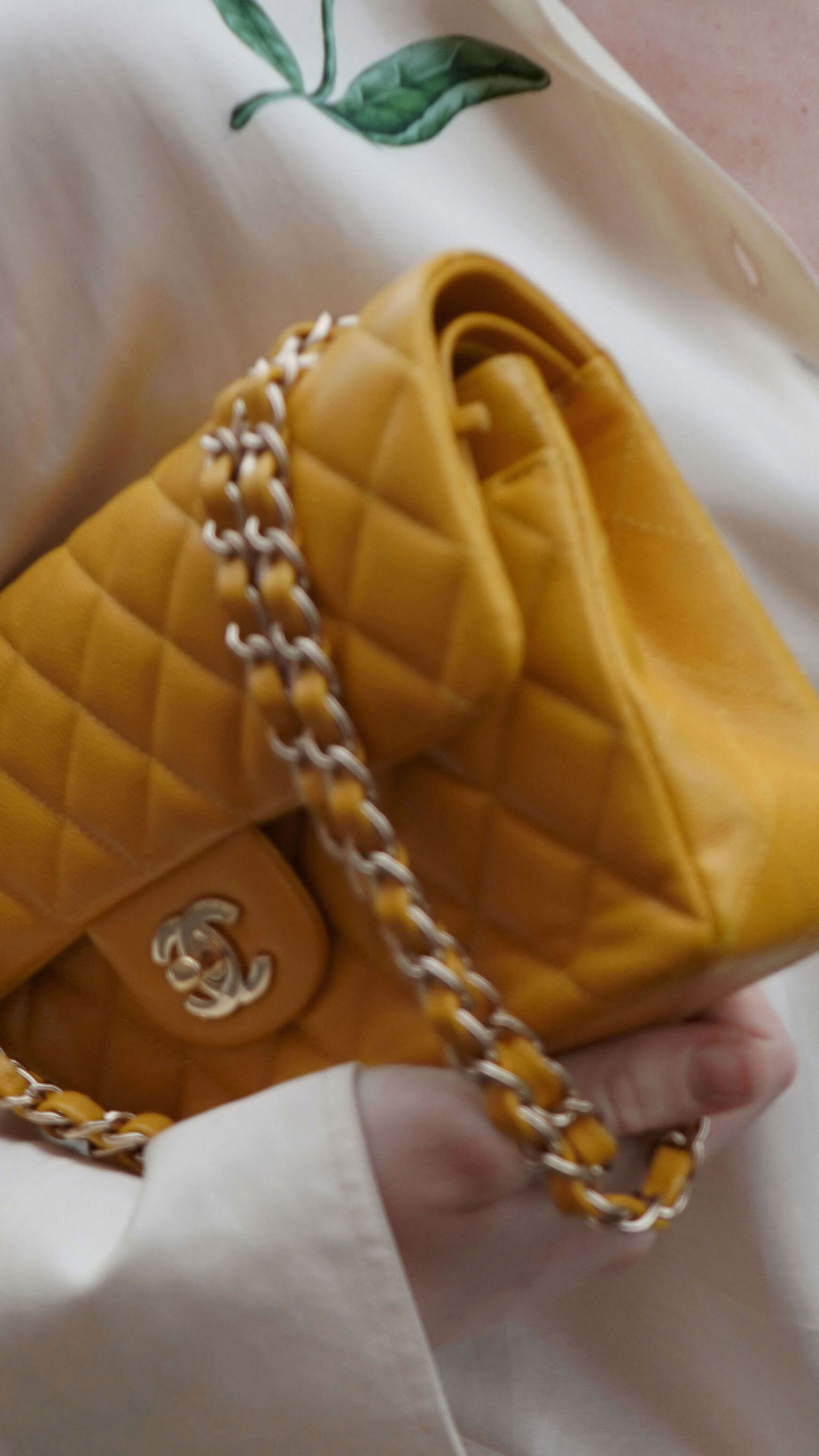 A side-by-side comparison image shows two yellow quilted Chanel handbags. On the left, a classic rectangular flap bag with gold-tone chain strap lies flat on a dark surface, featuring the iconic interlocking CC logo. On the right, a similar but more compact quilted bag with chain strap is held by a person wearing a white shirt, displayed against an outdoor urban background.