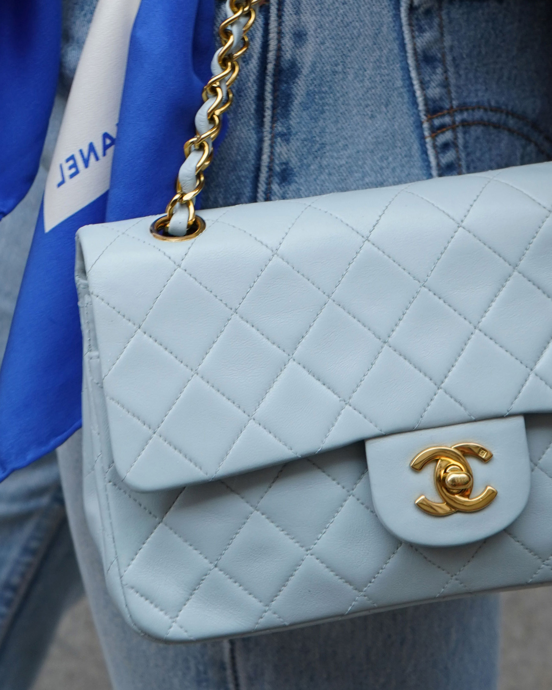 A close-up photo shows a light blue Chanel quilted leather handbag with gold chain strap, held by a hand against jeans, featuring a blue scarf with white Chanel logo draped over it.