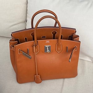 Person in beige coat and black heels crossing the street holding a beige and black Hermès bag.