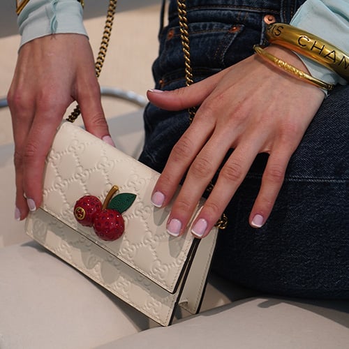A close-up of a Gucci Horsebit 1955 shoulder bag in GG Supreme canvas with brown leather trim and gold horsebit hardware, resting on a person's lap dressed in white clothing with light pink manicured nails.