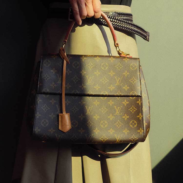 A Louis Vuitton Speedy 30 handbag in Damier Azur canvas with natural leather handles, adorned with a beige fur pom-pom and leather tassel charm, resting on a beige leather car seat.