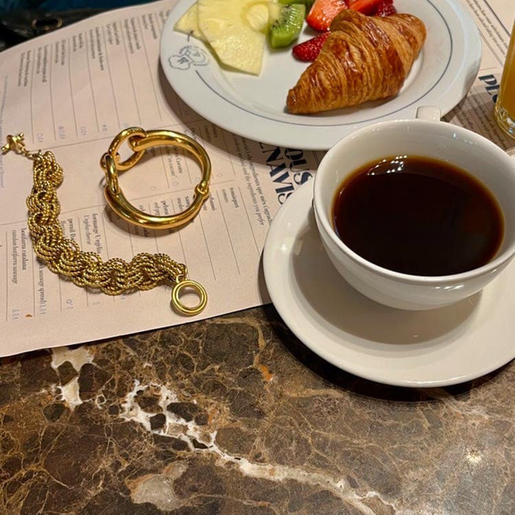 Breakfast table with black coffee, fruit, croissant, and two gold jewelry pieces on a menu.