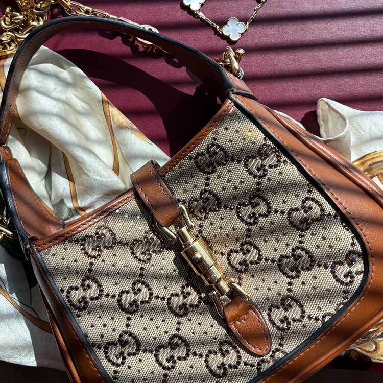 A close-up of a Gucci Horsebit 1955 shoulder bag in GG Supreme canvas with brown leather trim and gold horsebit hardware, resting on a person's lap dressed in white clothing with light pink manicured nails.
