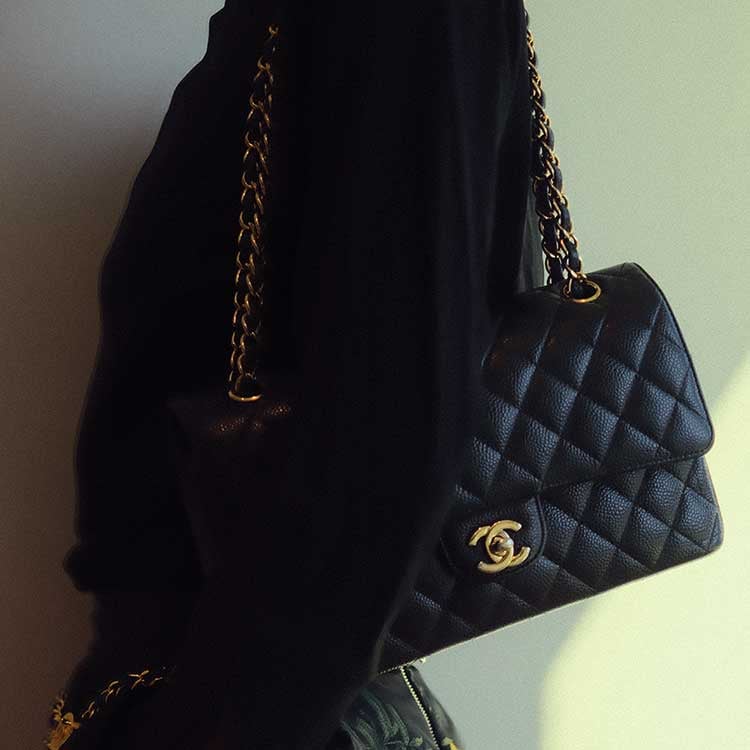 A hand holding a yellow Chanel quilted clutch bag in sunlight, featuring the signature CC turn-lock closure and a gold chain strap, with shadows cast on a light wall in the background.
