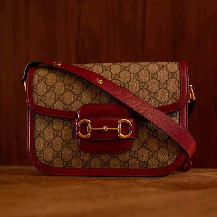 A close-up of a Gucci Horsebit 1955 shoulder bag in GG Supreme canvas with brown leather trim and gold horsebit hardware, resting on a person's lap dressed in white clothing with light pink manicured nails.