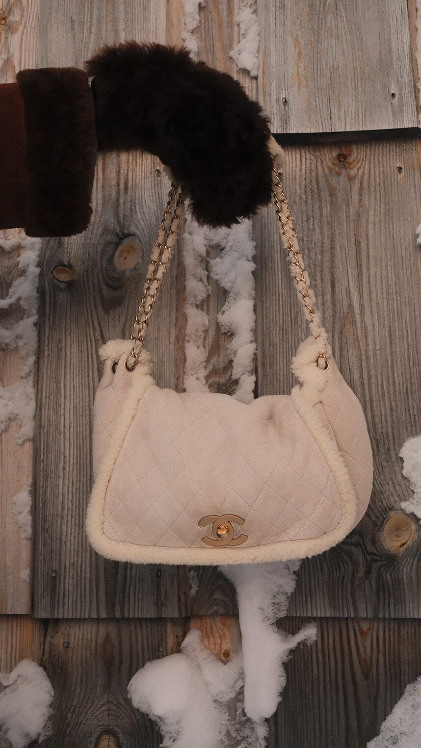 Flat lay of luxury accessories on a warm beige background, including gold jewelry, a black ribbon with decorative pins, a chain strap, a keyring with charms, and a small beige leather pouch.