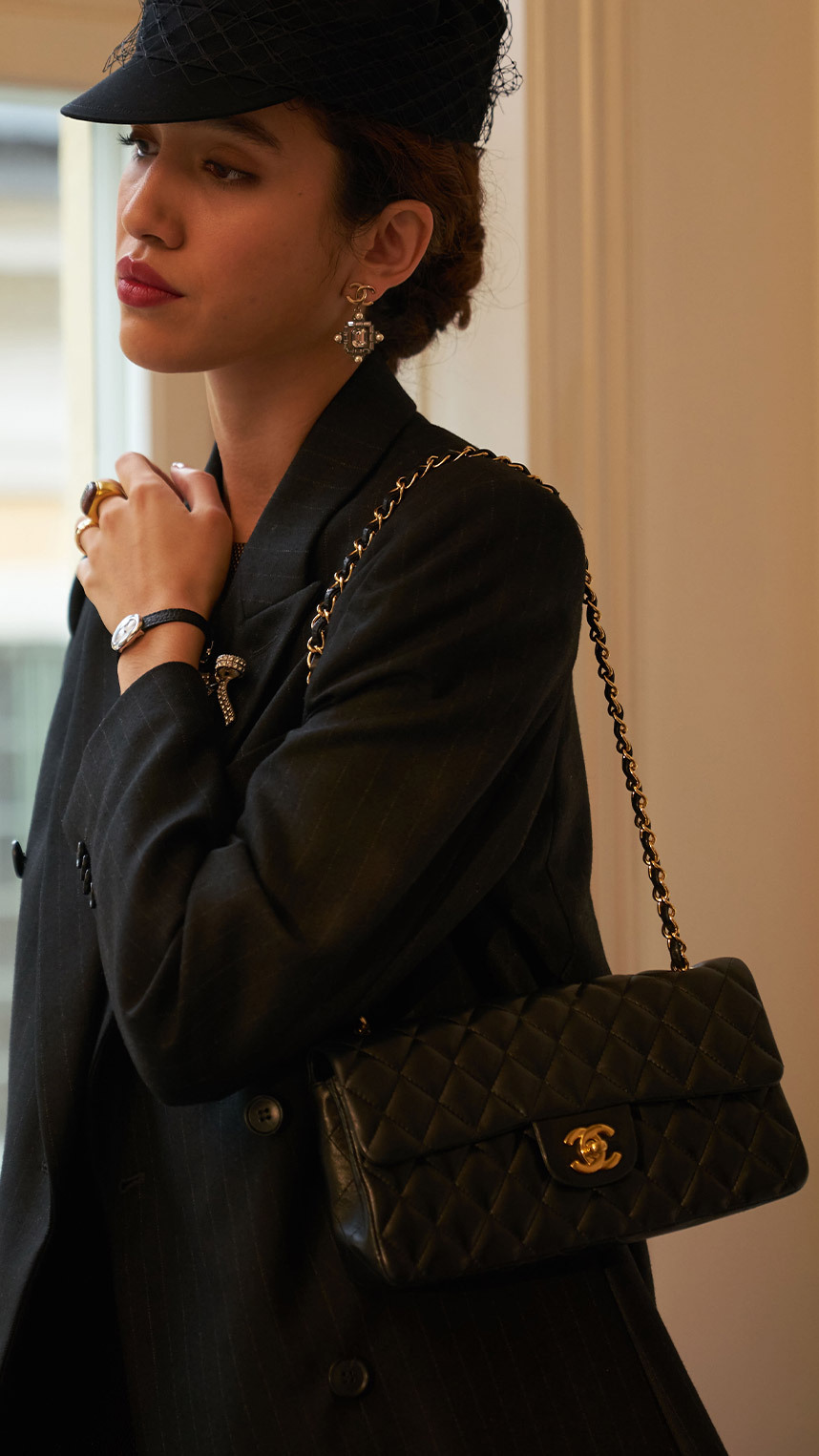Person dressed in elegant black attire, wearing a veiled hat, statement earrings, and decorative brooches, smiling while holding a quilted black handbag with gold hardware.