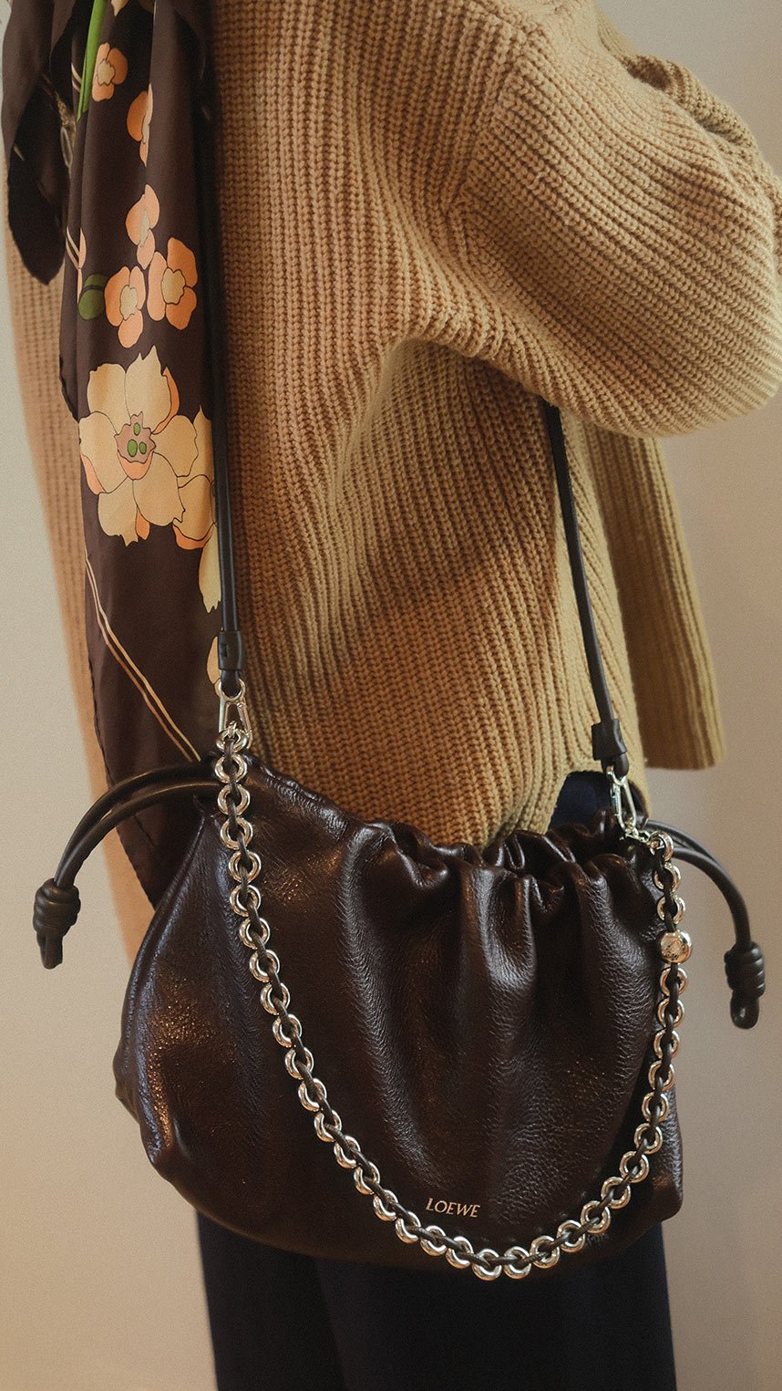 A person wearing a brown knit sweater carries a black Loewe leather shoulder bag with a silver chain detail.