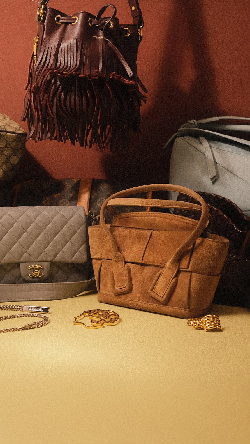 Person dressed in elegant black attire, wearing a veiled hat, statement earrings, and decorative brooches, smiling while holding a quilted black handbag with gold hardware.