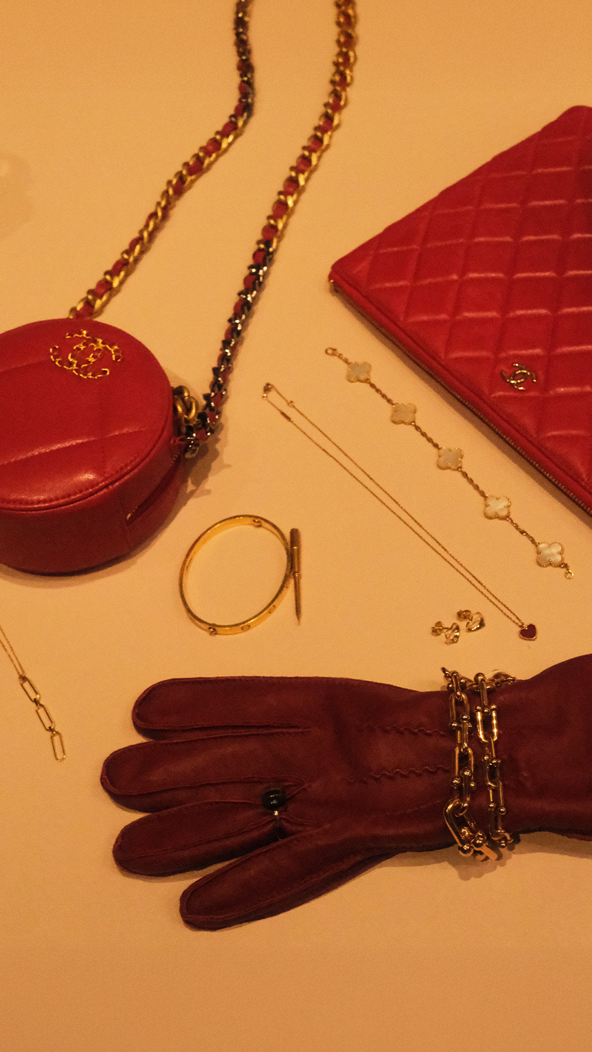 Flat lay of luxury accessories on a warm beige background, including gold jewelry, a black ribbon with decorative pins, a chain strap, a keyring with charms, and a small beige leather pouch.