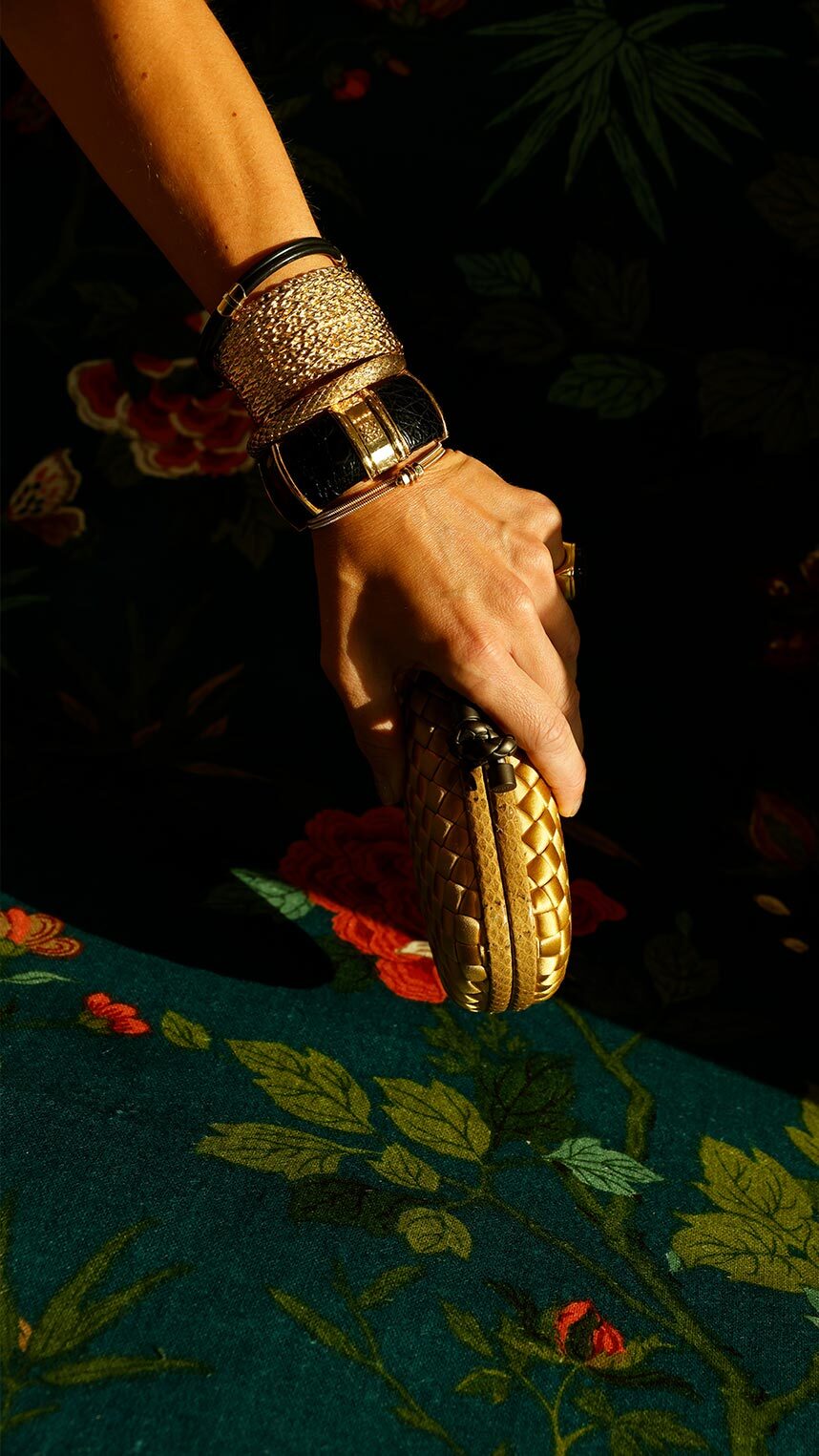 A close-up of a hand holding a small gold woven clutch, adorned with layered gold and black statement bracelets, against a dark floral background.