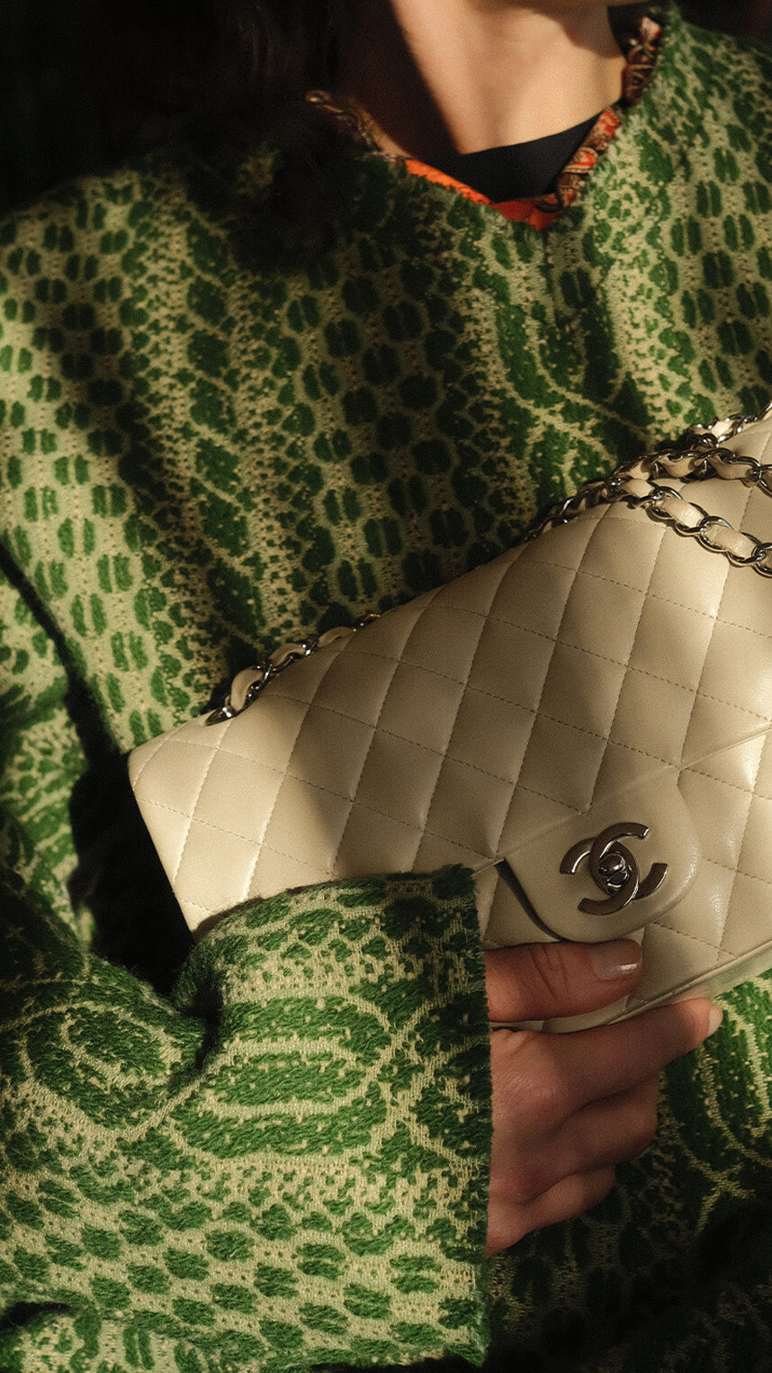 Flat lay of luxury accessories on a warm beige background, including gold jewelry, a black ribbon with decorative pins, a chain strap, a keyring with charms, and a small beige leather pouch.