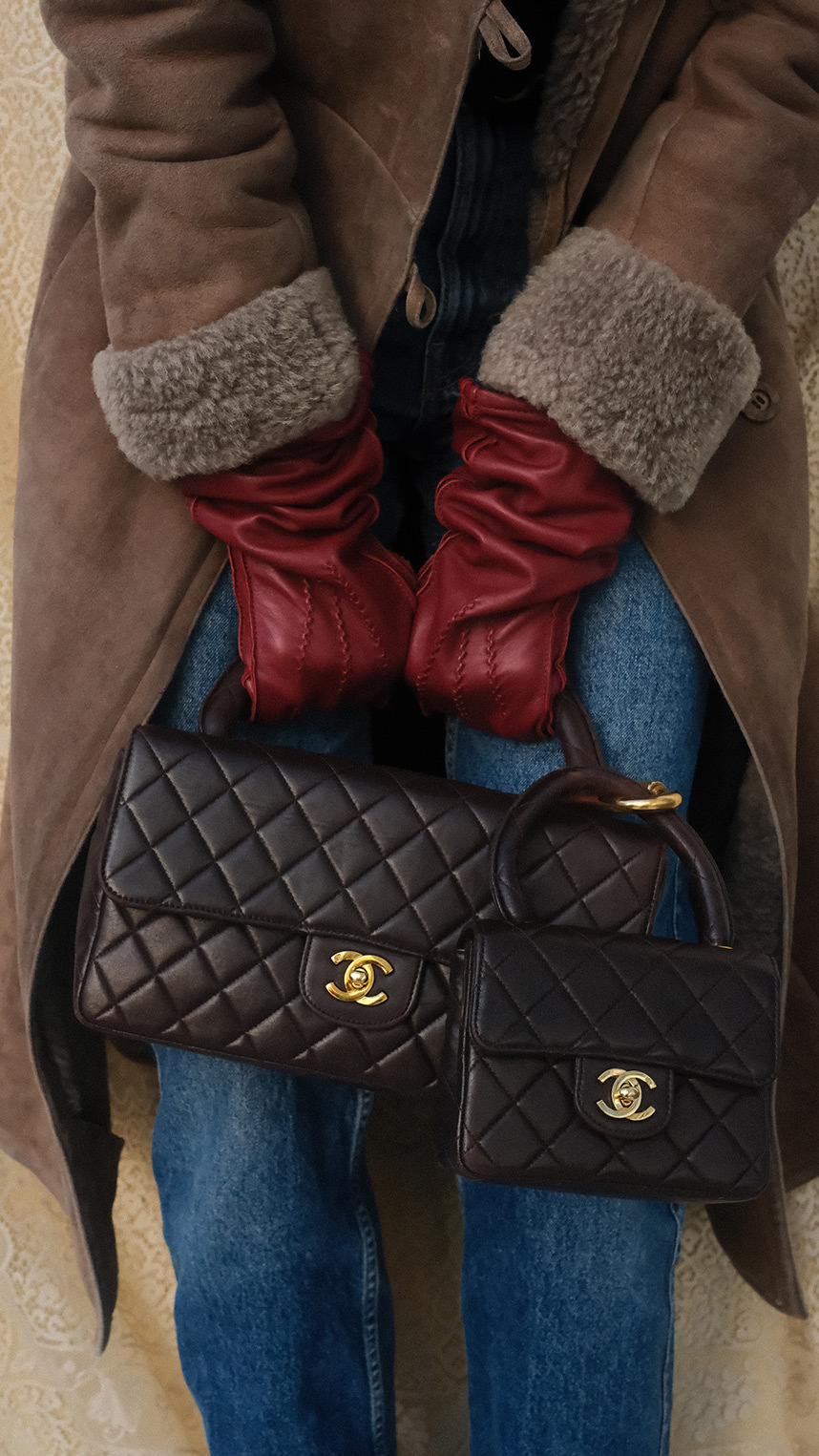 Flat lay of luxury accessories on a warm beige background, including gold jewelry, a black ribbon with decorative pins, a chain strap, a keyring with charms, and a small beige leather pouch.