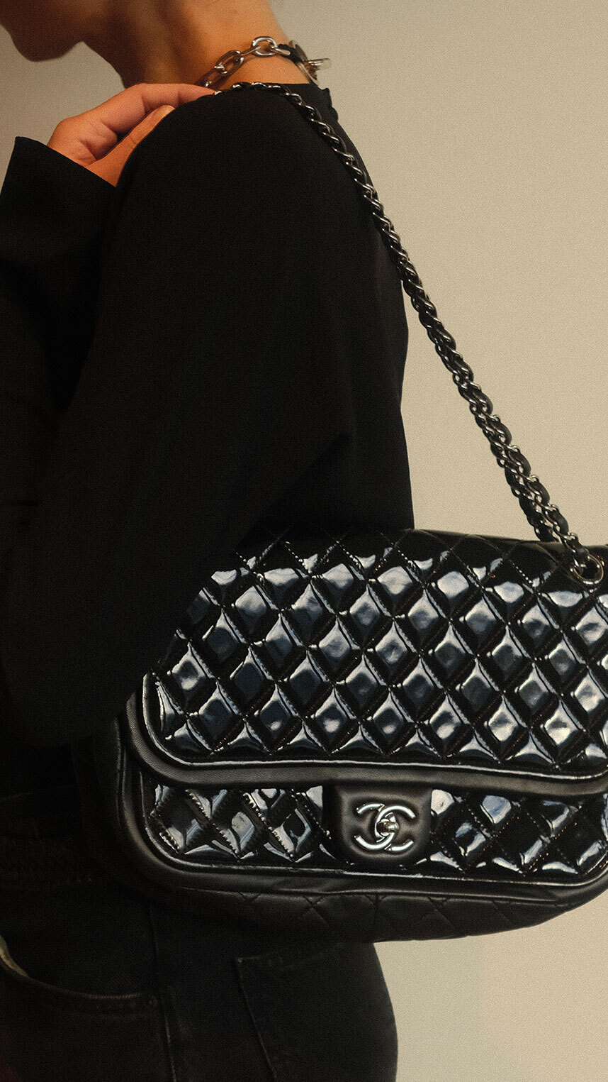 Person wearing a black outfit holding a black quilted Chanel shoulder bag with a chain strap and silver logo clasp against a neutral background.