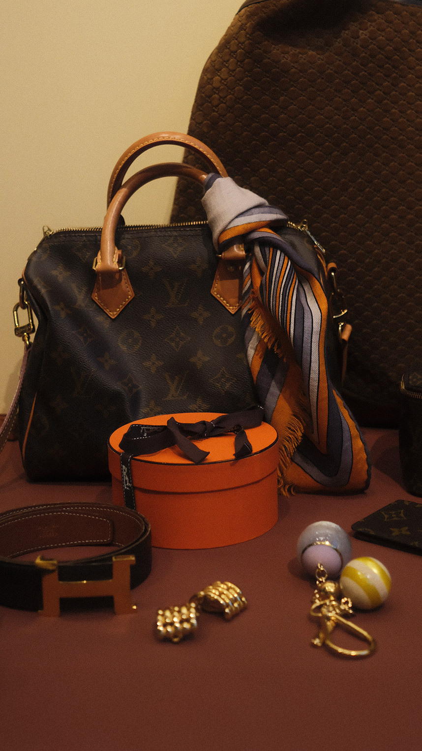 Person dressed in elegant black attire, wearing a veiled hat, statement earrings, and decorative brooches, smiling while holding a quilted black handbag with gold hardware.