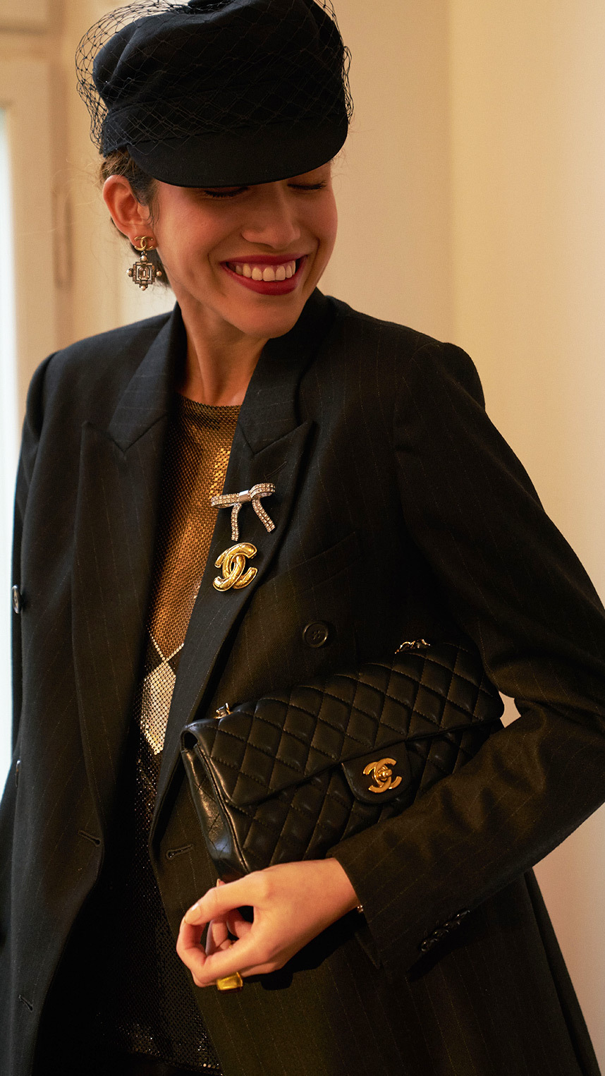 Person dressed in elegant black attire, wearing a veiled hat, statement earrings, and decorative brooches, smiling while holding a quilted black handbag with gold hardware.
