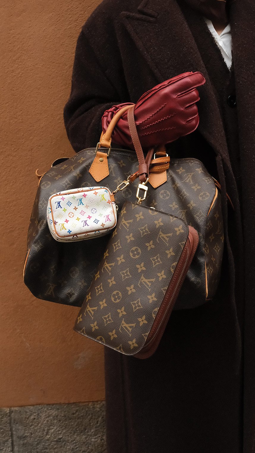 Flat lay of luxury accessories on a warm beige background, including gold jewelry, a black ribbon with decorative pins, a chain strap, a keyring with charms, and a small beige leather pouch.