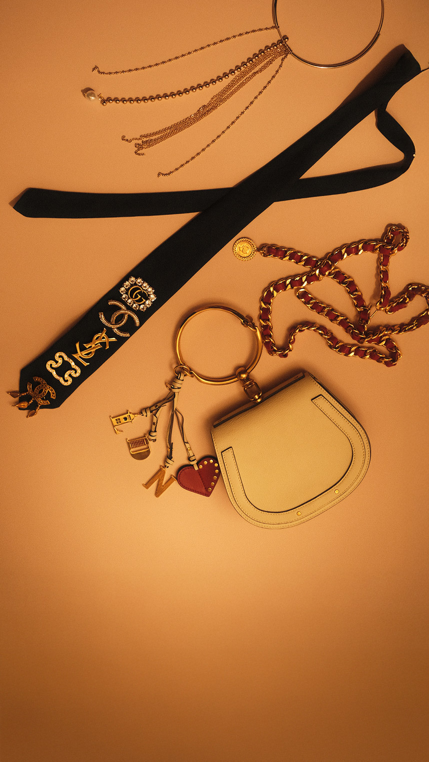 Flat lay of luxury accessories on a warm beige background, including gold jewelry, a black ribbon with decorative pins, a chain strap, a keyring with charms, and a small beige leather pouch.