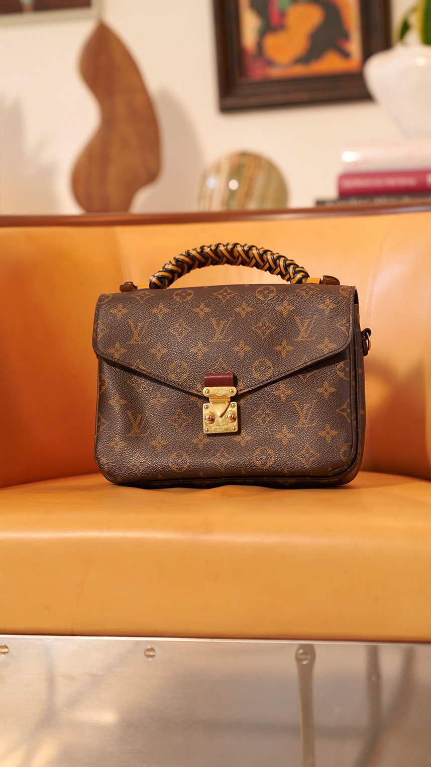 Person dressed in elegant black attire, wearing a veiled hat, statement earrings, and decorative brooches, smiling while holding a quilted black handbag with gold hardware.