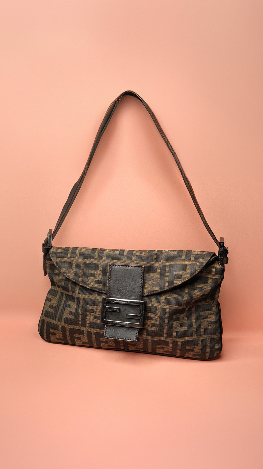 A Fendi Baguette handbag in brown Zucca canvas featuring the iconic FF monogram, a black leather flap with a metal logo buckle, and a single leather shoulder strap, displayed against a soft peach background.