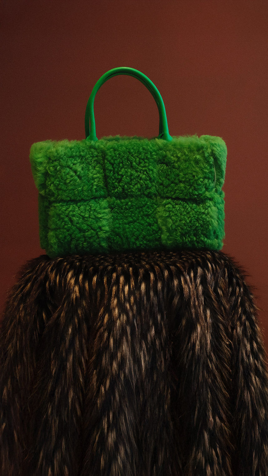 A person wearing a brown knit sweater carries a black Loewe leather shoulder bag with a silver chain detail.