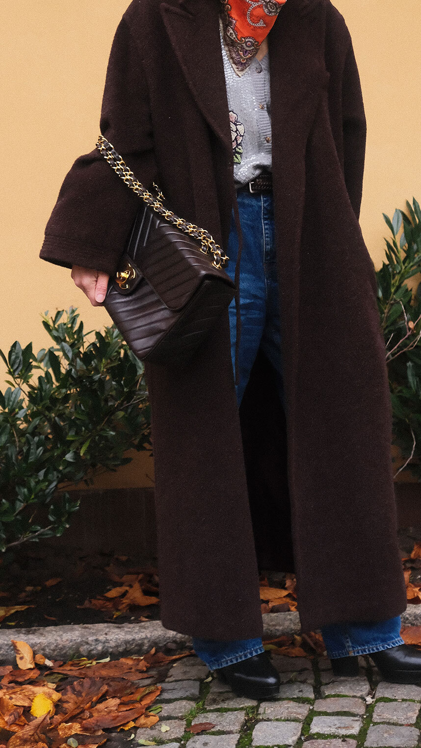 Person dressed in elegant black attire, wearing a veiled hat, statement earrings, and decorative brooches, smiling while holding a quilted black handbag with gold hardware.