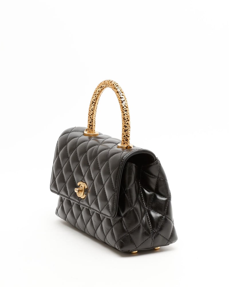 Angled profile of a Chanel Small Signature Coco Handle Bag in black quilted leather, showing its ornate gold-tone handle.