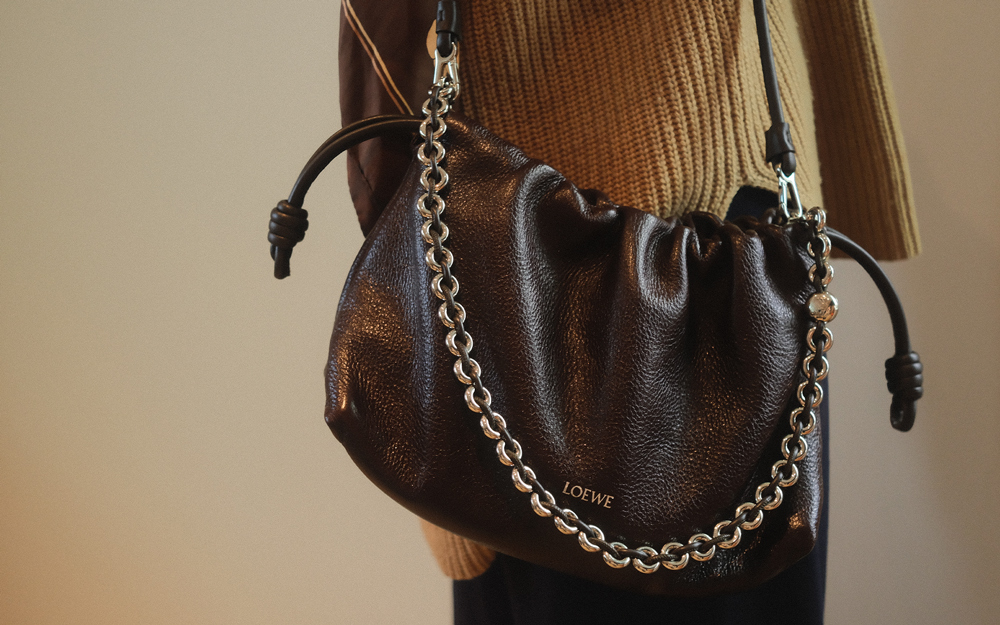 A person wearing a brown knit sweater carries a black Loewe leather shoulder bag with a silver chain detail.