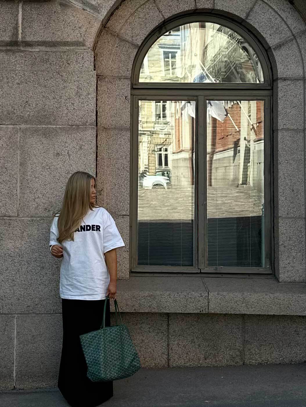 Woman with Goyard bag in white T-shirt and black skirt by stone wall with arched window.