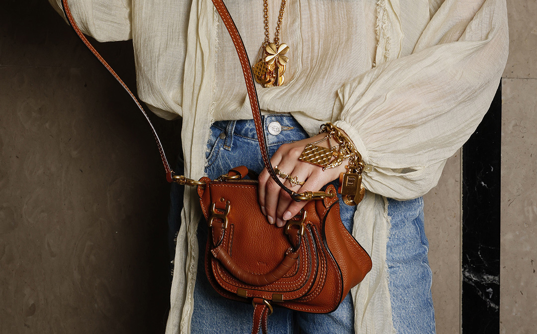 Close-up of a person wearing a sheer light blouse and blue jeans, styled with a small brown leather crossbody bag featuring gold hardware. The look is completed with layered gold jewelry – including necklaces, bracelets, and rings – evoking a luxurious boho-chic vibe.