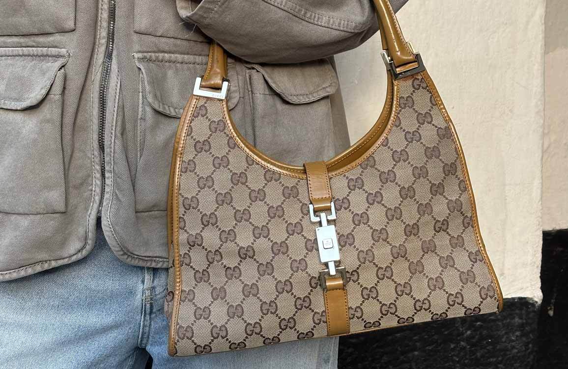 Person wearing brown trousers holding a beige Gucci bag with brown trim.
