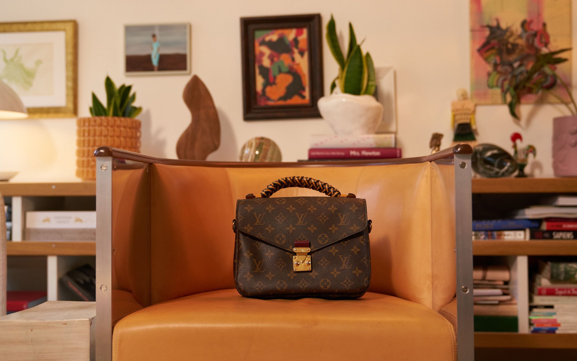 A person dressed in an elegant black outfit and veiled hat, accessorized with statement earrings and brooches. In the first view, they smile while holding a quilted black handbag with gold hardware. In the second view, they stand by a window, reaching toward the handbag resting on a stack of books.