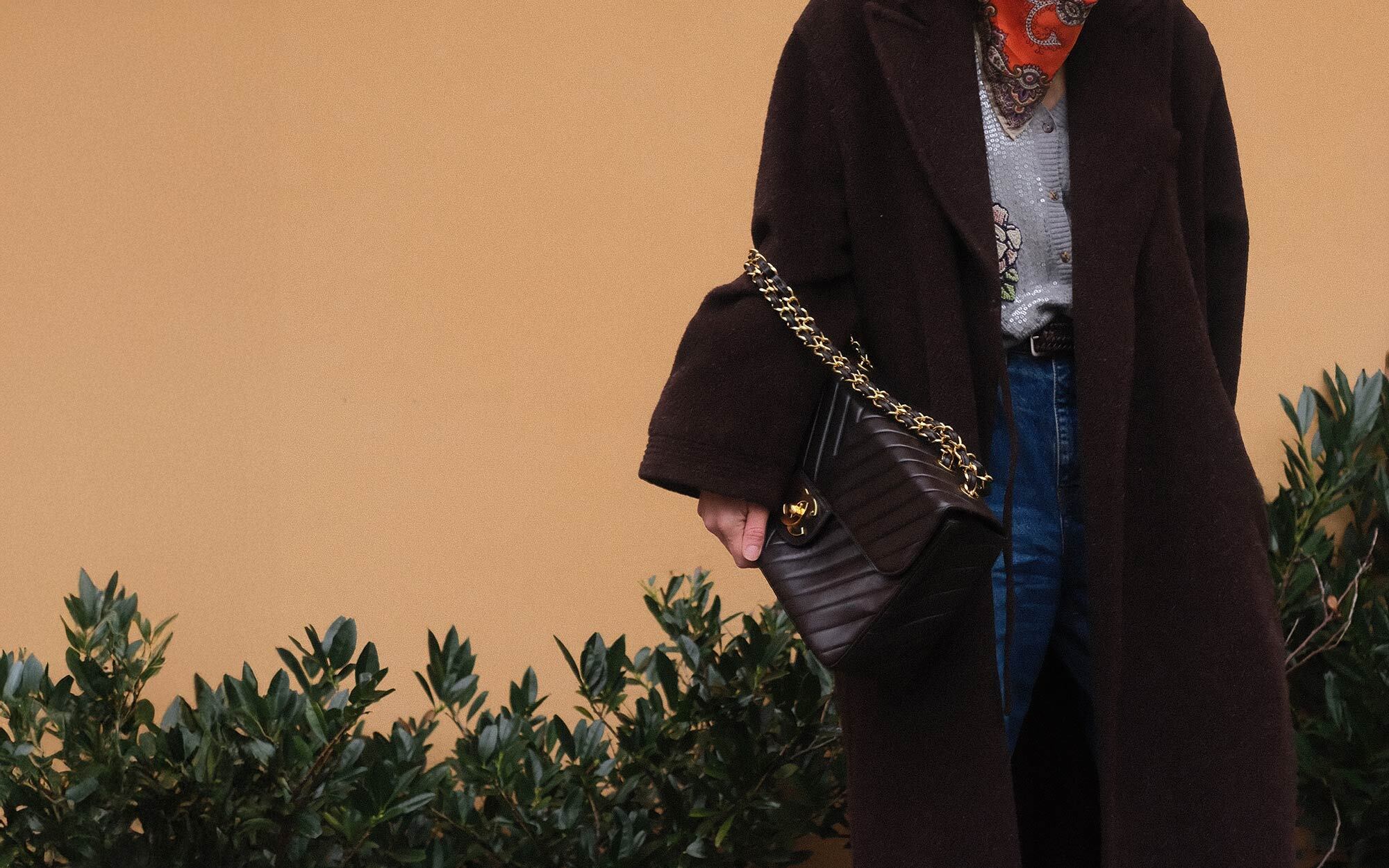A person dressed in an elegant black outfit and veiled hat, accessorized with statement earrings and brooches. In the first view, they smile while holding a quilted black handbag with gold hardware. In the second view, they stand by a window, reaching toward the handbag resting on a stack of books.
