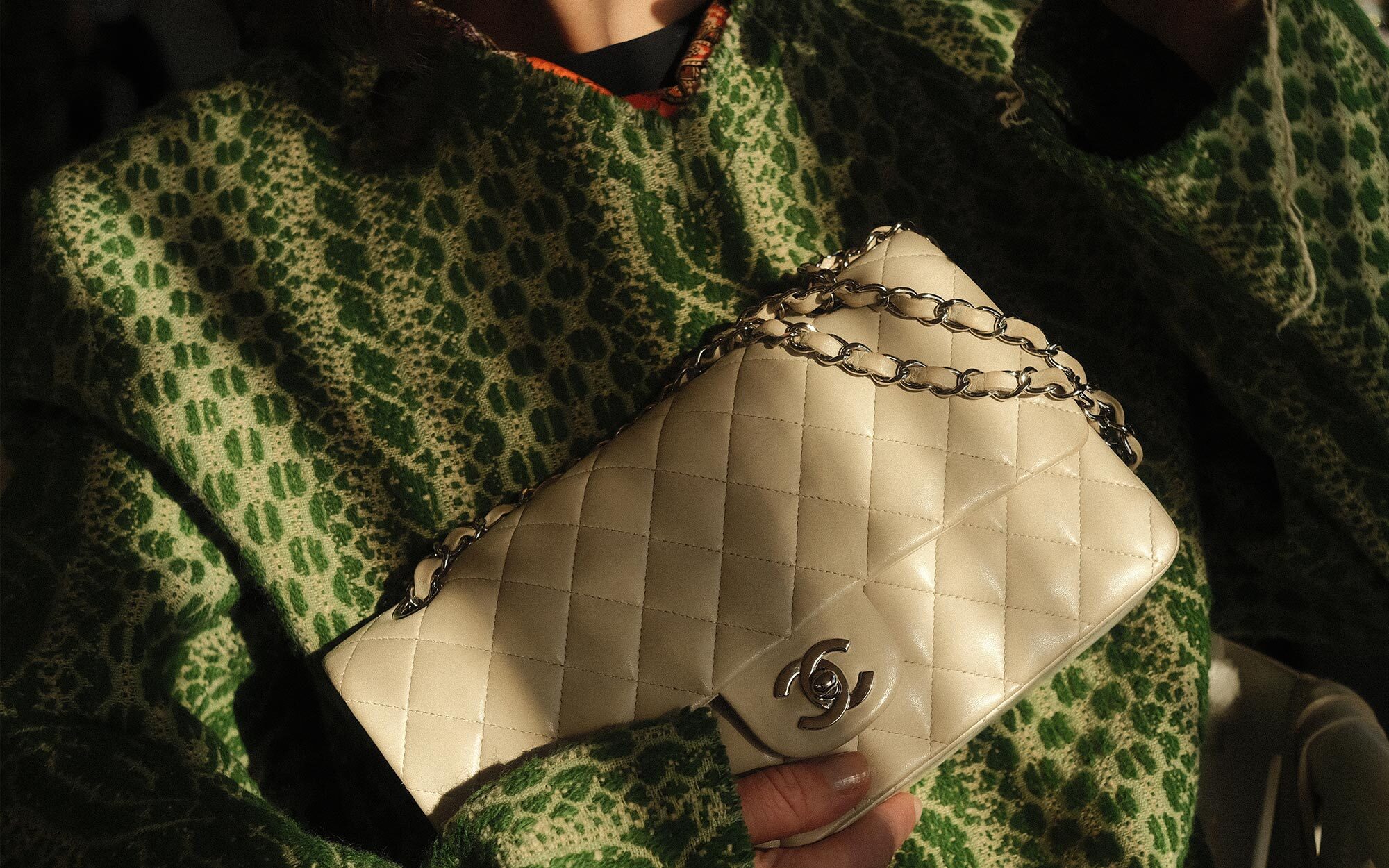 A person dressed in an elegant black outfit and veiled hat, accessorized with statement earrings and brooches. In the first view, they smile while holding a quilted black handbag with gold hardware. In the second view, they stand by a window, reaching toward the handbag resting on a stack of books.