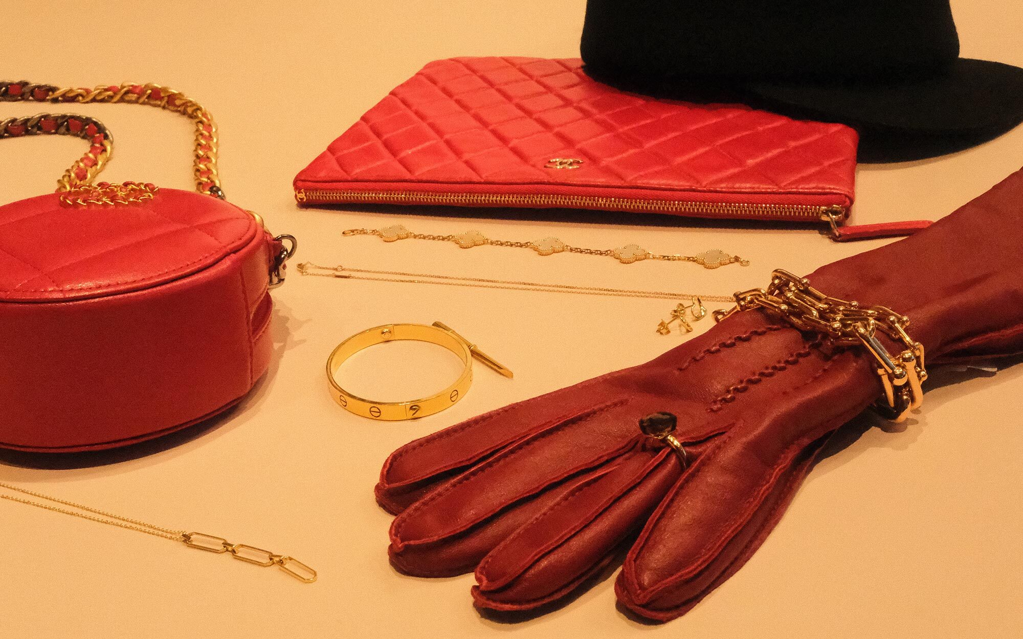 A person dressed in an elegant black outfit and veiled hat, accessorized with statement earrings and brooches. In the first view, they smile while holding a quilted black handbag with gold hardware. In the second view, they stand by a window, reaching toward the handbag resting on a stack of books.