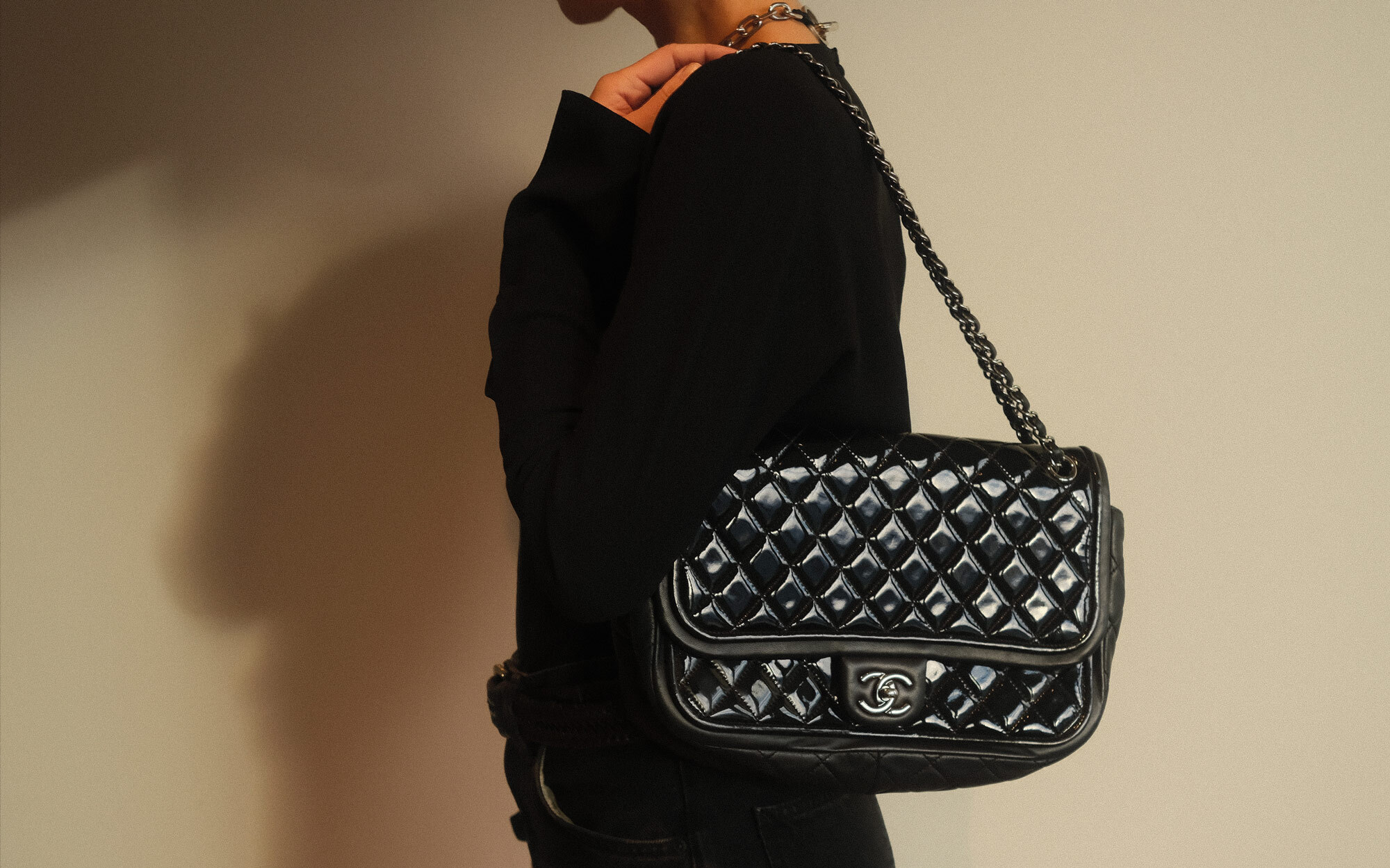 ALT text: Person wearing a black outfit holding a black quilted Chanel shoulder bag with a chain strap and silver logo clasp against a neutral background.