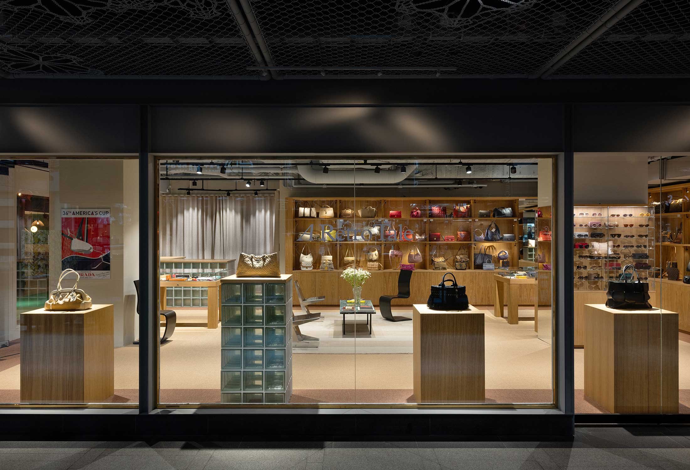 A luxury boutique interior featuring wooden shelves lined with designer handbags, including Hermès, Chanel, and Saint Laurent, with a black modern chair and a table decorated with books and a floral arrangement in the foreground.