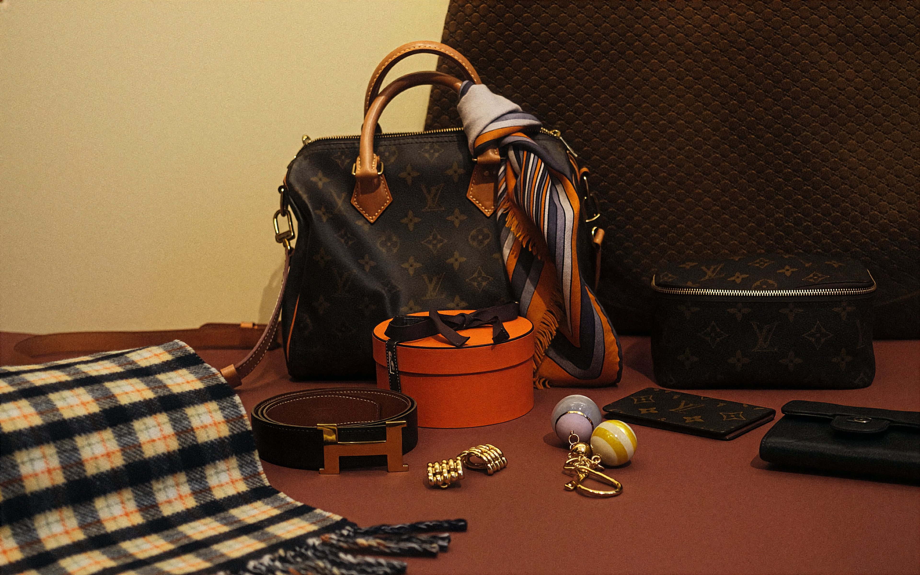 A person dressed in an elegant black outfit and veiled hat, accessorized with statement earrings and brooches. In the first view, they smile while holding a quilted black handbag with gold hardware. In the second view, they stand by a window, reaching toward the handbag resting on a stack of books.