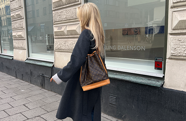 Woman walking on a city street wearing a dark coat and carrying a brown Louis Vuitton monogram bucket bag.