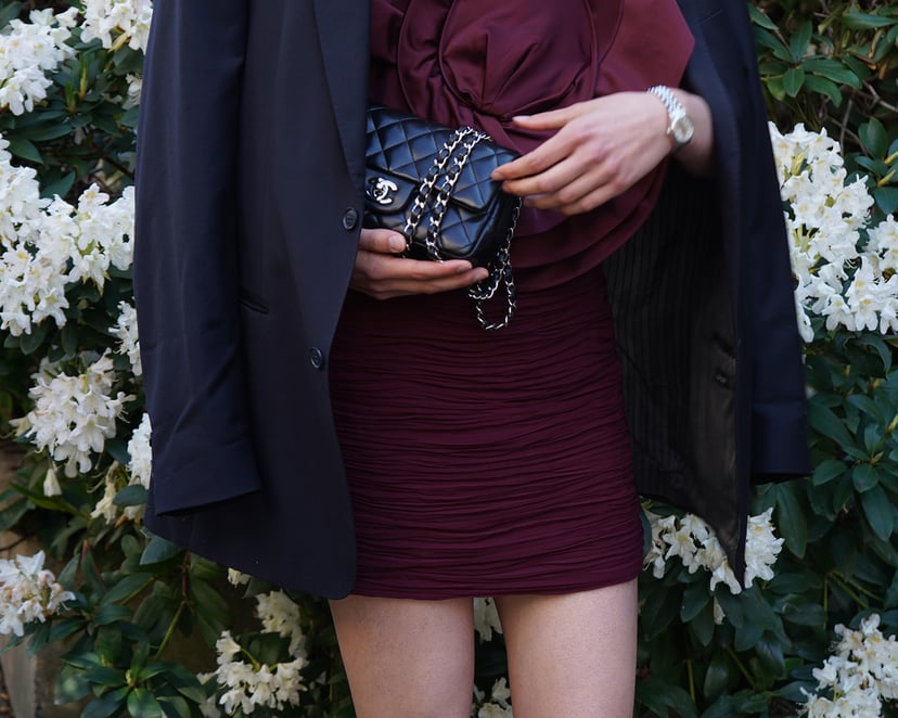 Close-up of a person wearing a sheer light blouse and blue jeans, styled with a small brown leather crossbody bag featuring gold hardware. The look is completed with layered gold jewelry – including necklaces, bracelets, and rings – evoking a luxurious boho-chic vibe.