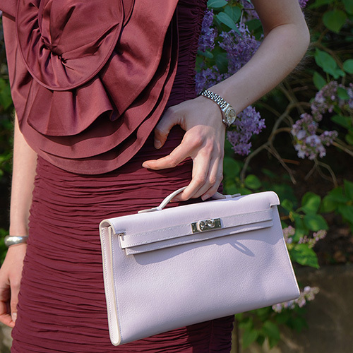 Close-up of a person wearing a sheer light blouse and blue jeans, styled with a small brown leather crossbody bag featuring gold hardware. The look is completed with layered gold jewelry – including necklaces, bracelets, and rings – evoking a luxurious boho-chic vibe.