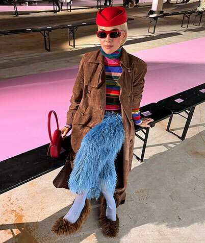 Woman in light jacket and sunglasses holding a brown Louis Vuitton bag with a patterned scarf.