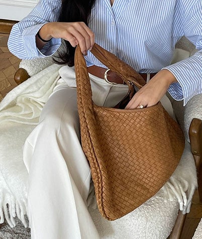 Person in blue and white shirt sitting with a large brown woven Bottega Veneta bag on their lap.