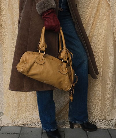 A stylish woman with long blonde hair, wearing sunglasses and a brown outfit, stands at a Parisian-style newsstand reading a magazine. She carries a textured brown shoulder bag, while the stand around her is filled with colorful newspapers and magazines.
