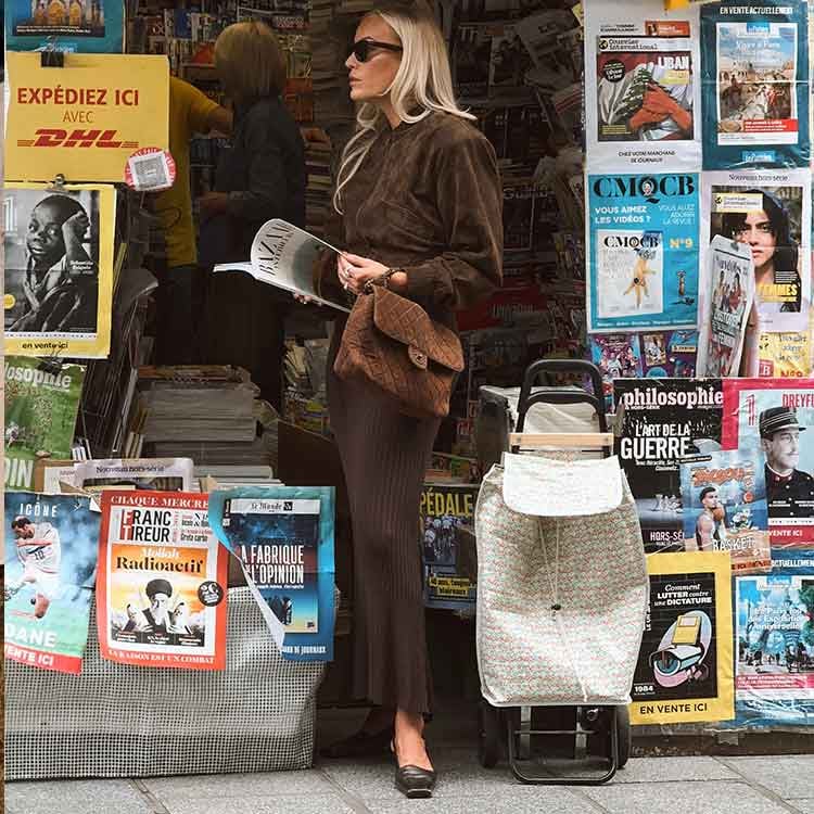 A stylish woman with long blonde hair, wearing sunglasses and a brown outfit, stands at a Parisian-style newsstand reading a magazine. She carries a textured brown shoulder bag, while the stand around her is filled with colorful newspapers and magazines.