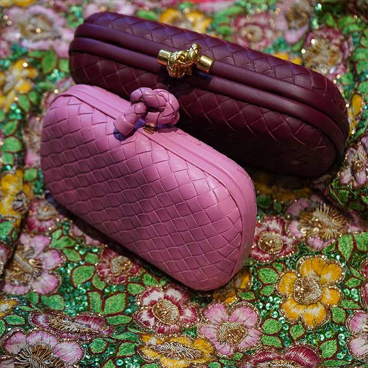 Close-up of a person wearing a sheer light blouse and blue jeans, styled with a small brown leather crossbody bag featuring gold hardware. The look is completed with layered gold jewelry – including necklaces, bracelets, and rings – evoking a luxurious boho-chic vibe.