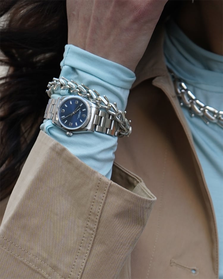 A stylish woman with long blonde hair, wearing sunglasses and a brown outfit, stands at a Parisian-style newsstand reading a magazine. She carries a textured brown shoulder bag, while the stand around her is filled with colorful newspapers and magazines.