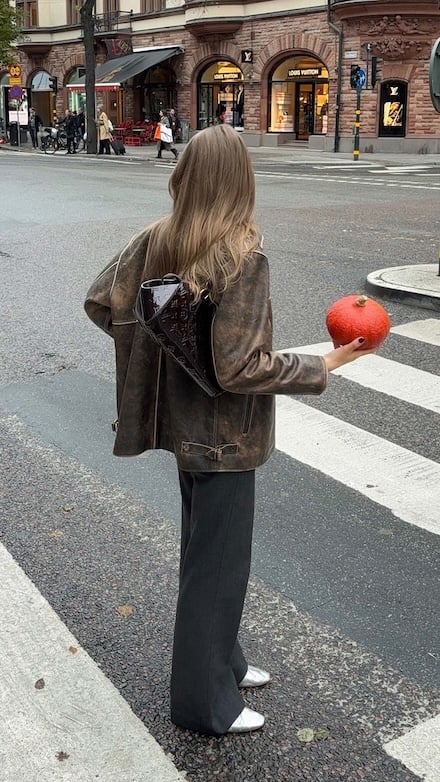 Woman in brown jacket with Louis Vuitton bag holding a fruit at a crosswalk.